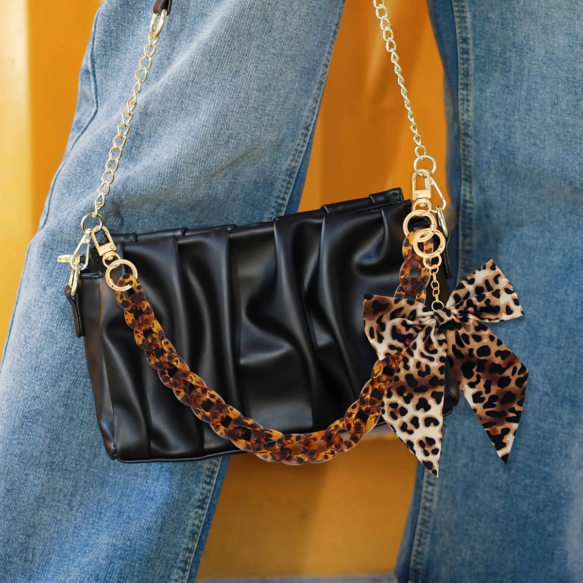 Schwarze Kunstleder-Tasche mit Leopardenmuster-Riemen und Schleifchen-Anhänger an blauen Jeans.