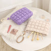 A purple quilted puffer makeup bag rests on a white fluffy rug next to cosmetic brushes.
