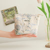 A hand holds a light blue floral canvas makeup bag with a small tag against a white background.
