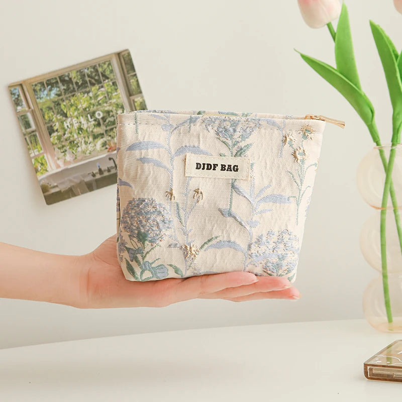 A hand holds a light blue floral canvas makeup bag with a small tag against a white background.