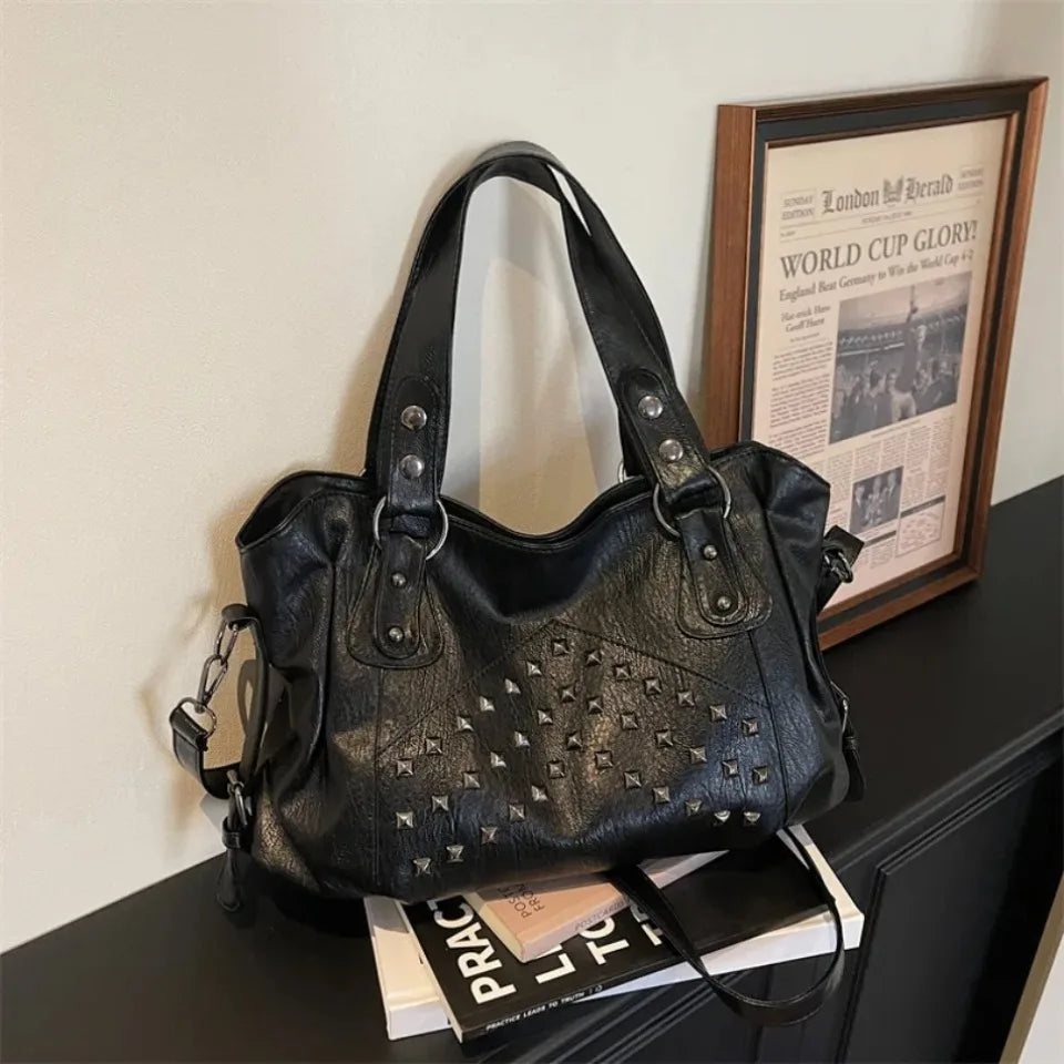 A black punk rivet leather tote bag resting on a dark surface next to a framed poster.
