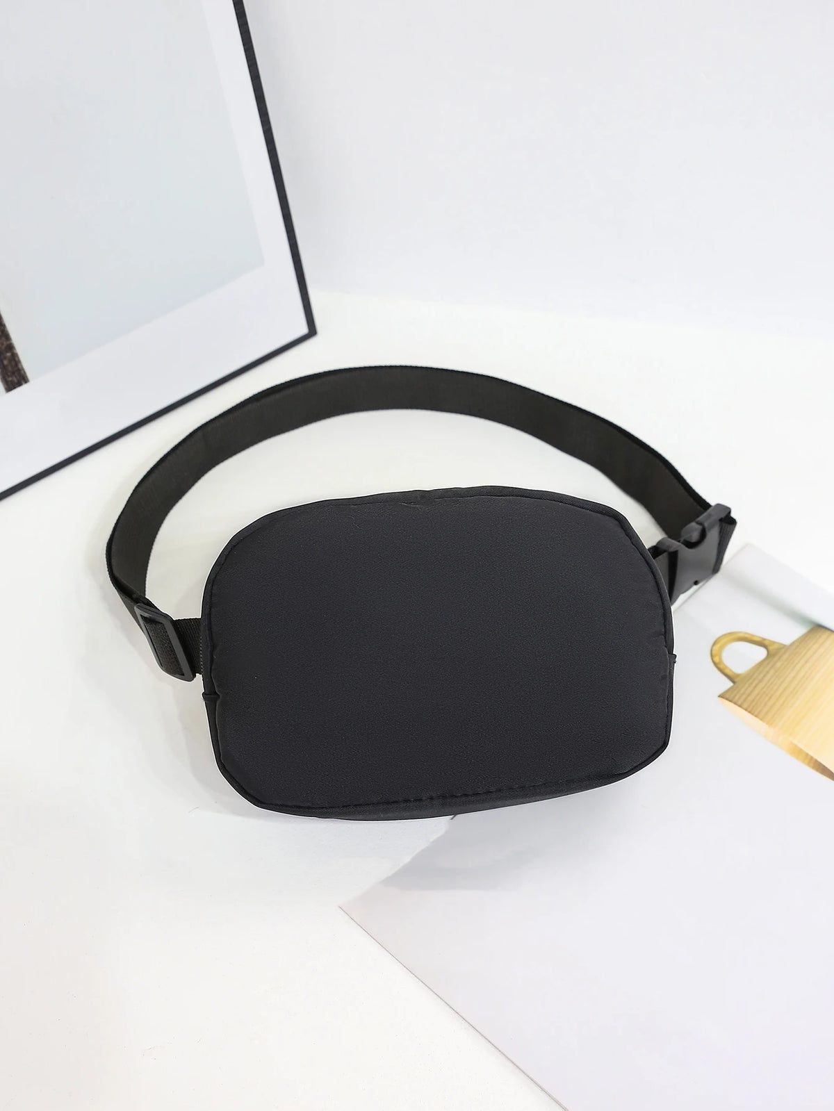 A black nylon cross-body waist bag is shown resting on a white surface next to a piece of paper.