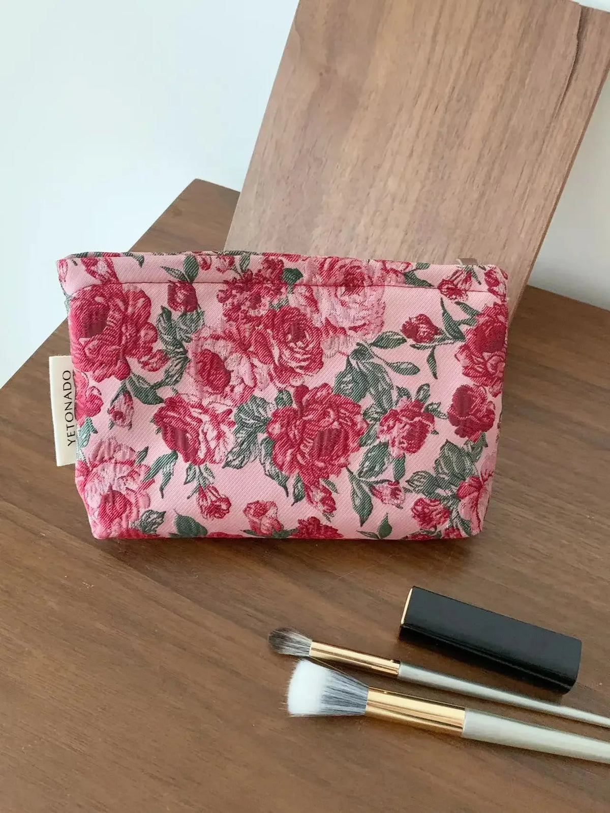 A pink floral cosmetic pouch with a red rose pattern rests on a wooden surface next to a makeup brush.