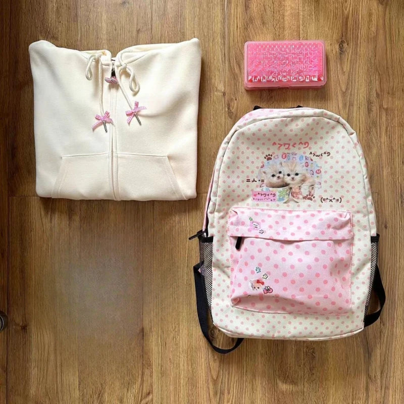 Sac à dos de chat rose et blanc à pois, à côté d'un sweat beige clair et d'un objet rectangulaire rose sur une surface en boi
