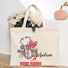 A beige canvas tote bag featuring a pink heart, stethoscope, and the name Madison with space for a personalized name.
