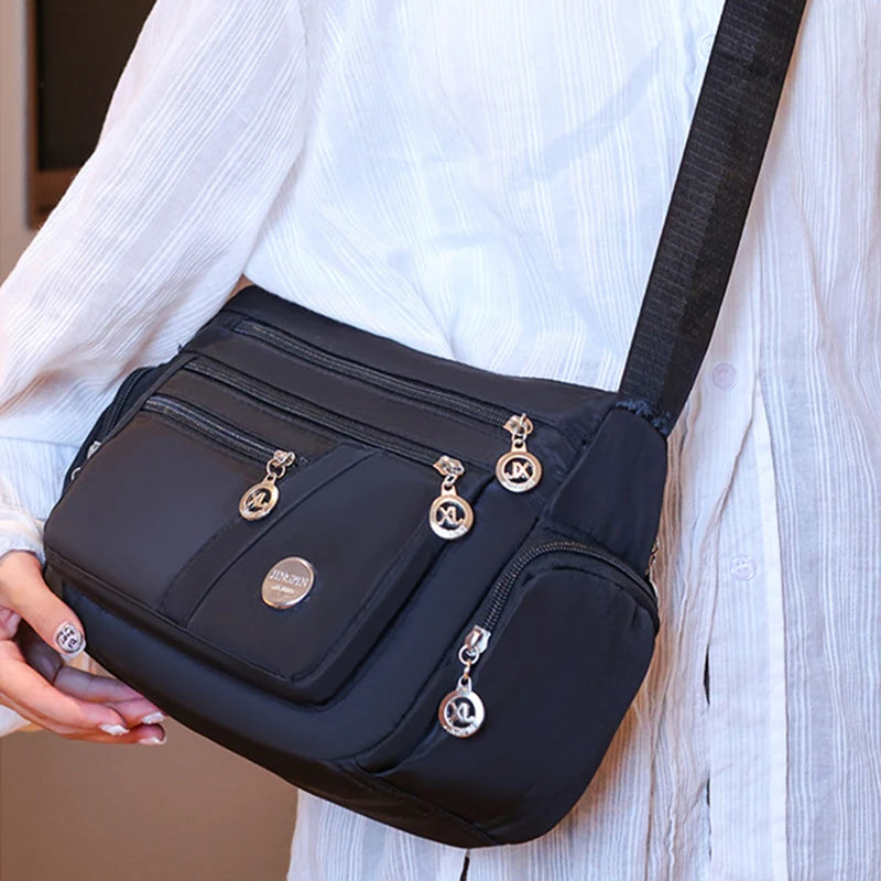 A black waterproof nylon crossbody tote bag with gold hardware is being held by a person wearing a white shirt.