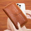 A brown PU leather zipper clutch wallet is held by a hand next to a white smartphone.