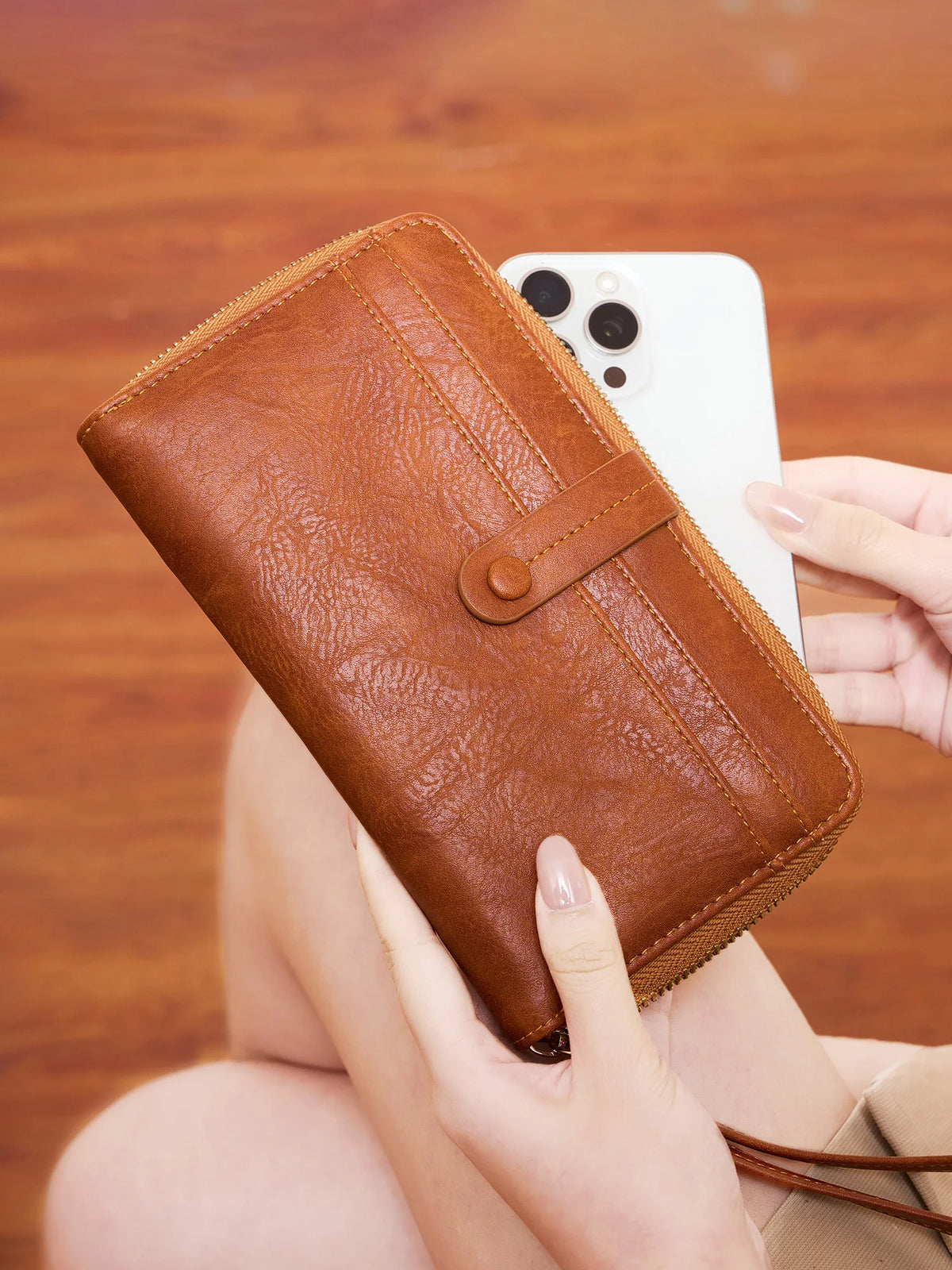 A brown PU leather zipper clutch wallet is held by a hand next to a white smartphone.