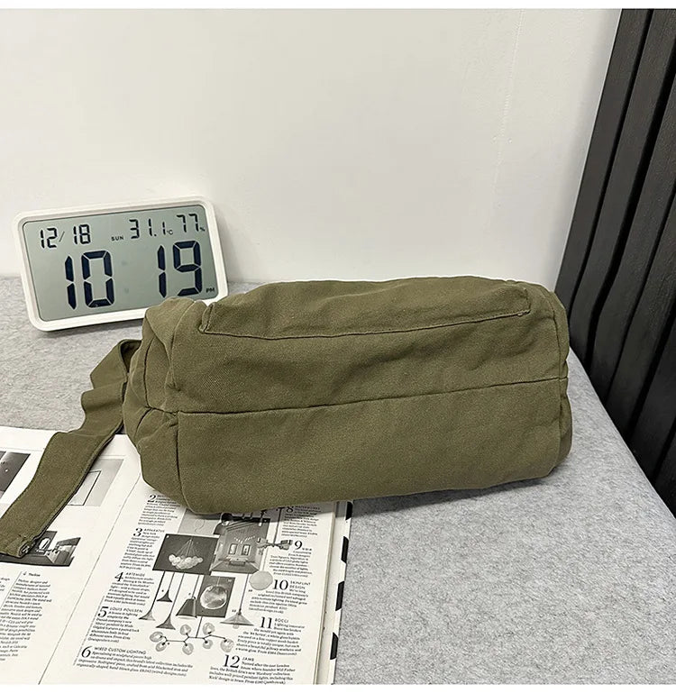 A olive green cotton canvas shoulder crossbody bag resting on a gray surface next to a digital clock and some printed materia