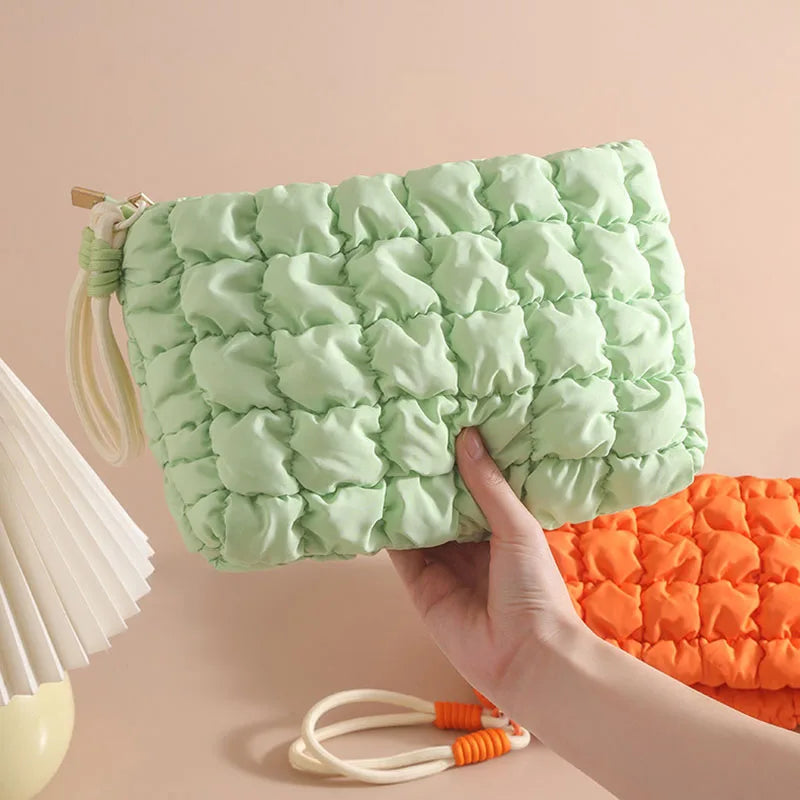 A hand holds a light green quilted puffer makeup bag against a beige background.