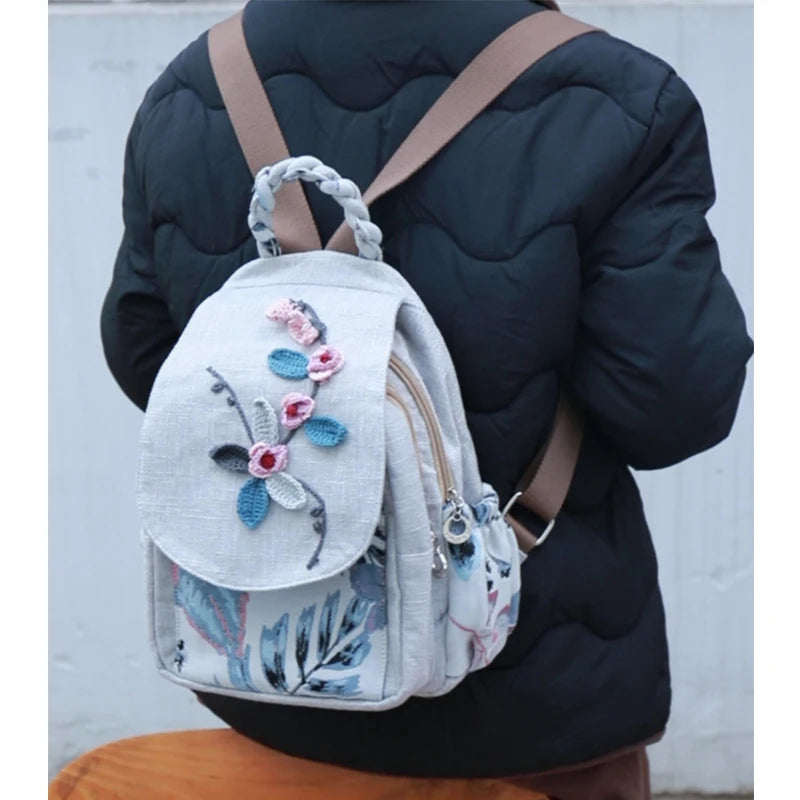 A light gray linen backpack with floral embroidery is worn by a person in a black quilted jacket.