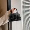 A hand holds a black leather square crossbody bag with a silver chain strap against a white window blind.