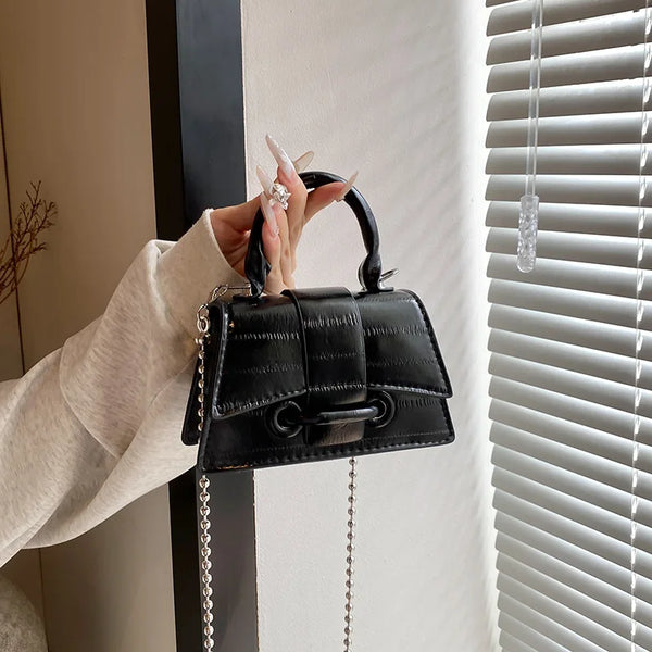 A hand holds a black leather square crossbody bag with a silver chain strap against a white window blind.