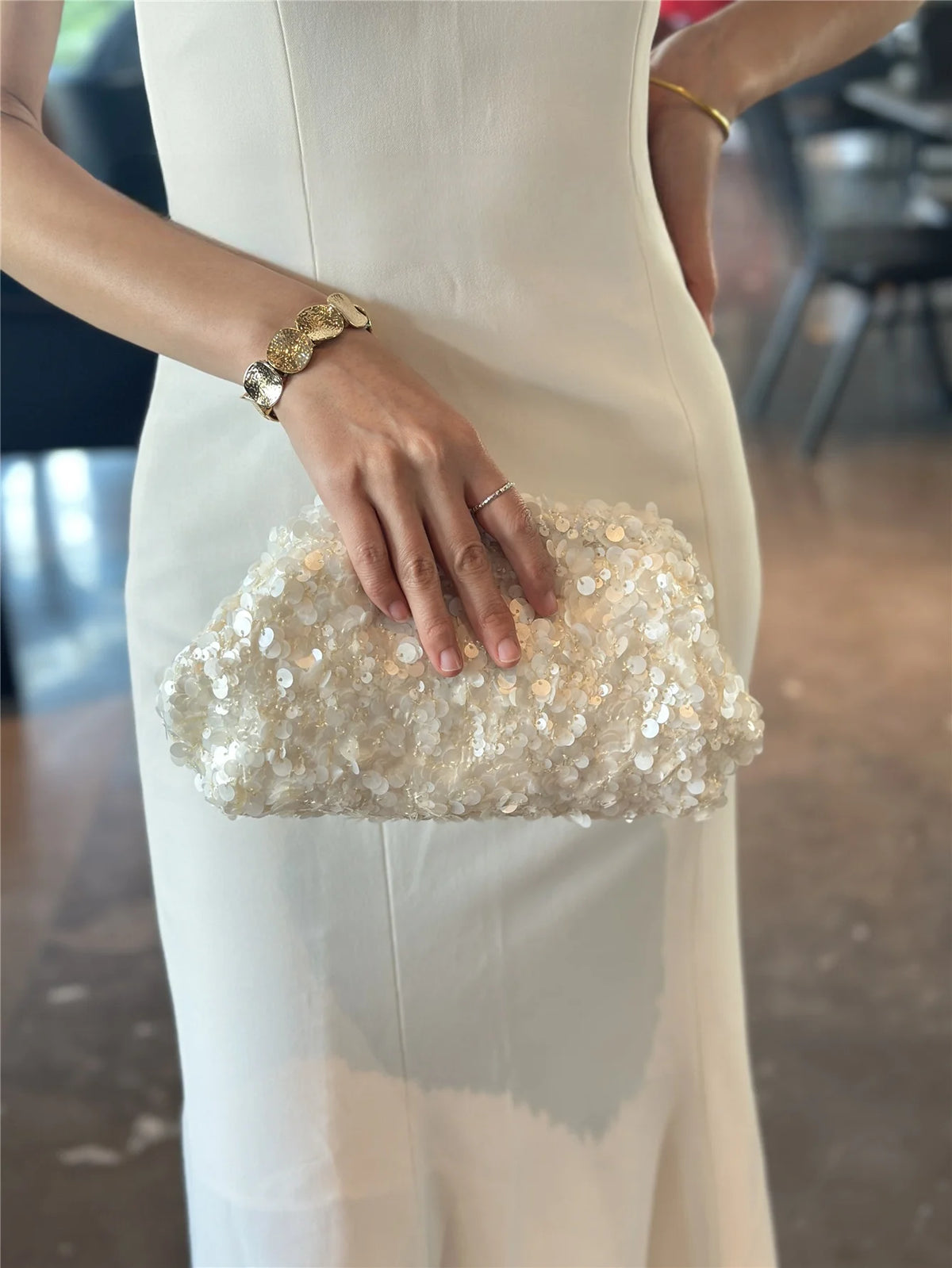 A woman wearing a white dress holds a light gold beaded chain shoulder clutch bag.
