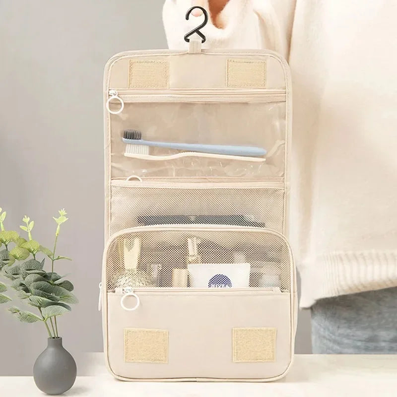 A beige waterproof makeup organizer pouch with clear compartments and a brush inside, held by a person's hand.