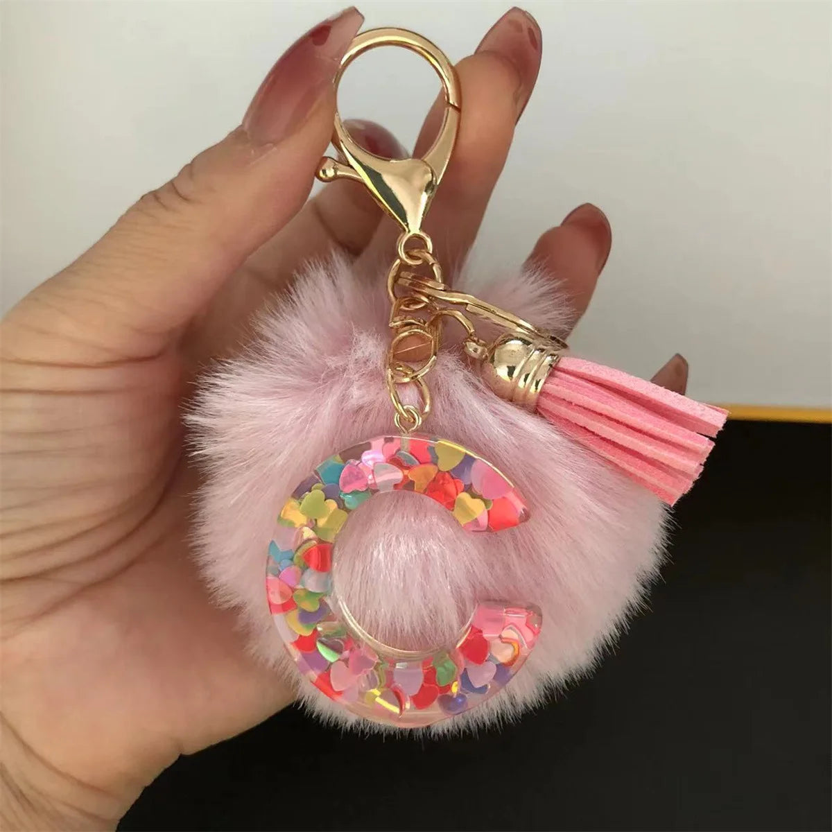 A gold-colored keychain featuring a resin initial letter charm with pink alphabet charms attached to a fluffy pink pom-pom.