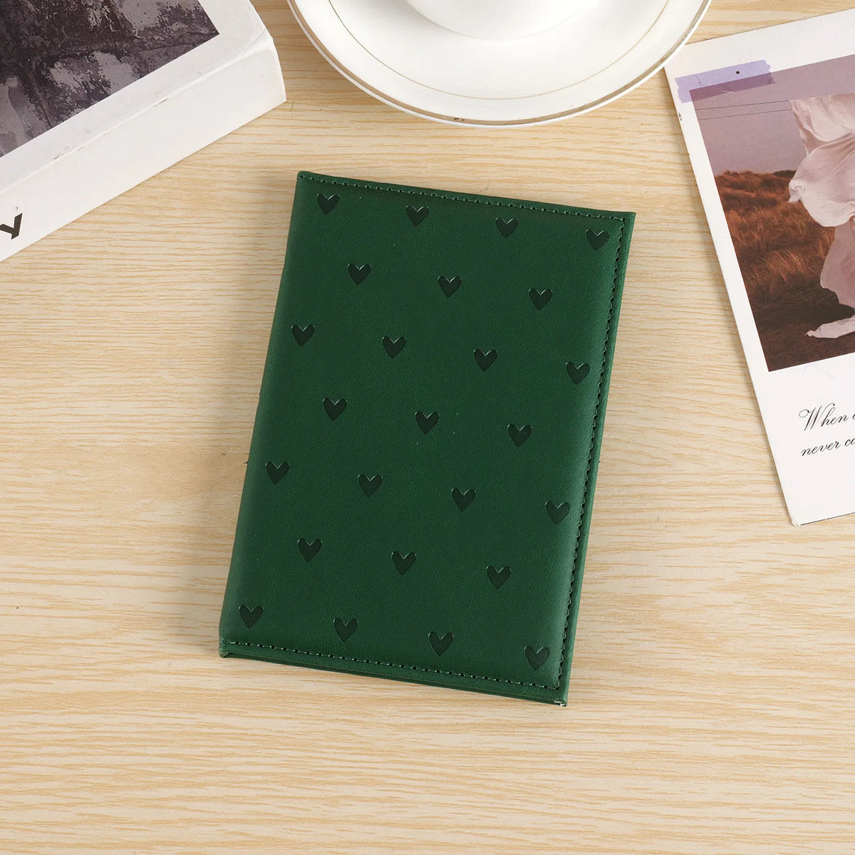 A dark green heart print passport cover travel wallet resting on a light wooden surface next to a white plate and a photo.