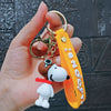 A hand holds a yellow silicone keychain featuring a white Snoopy dog with a brown floppy ear and a red collar.