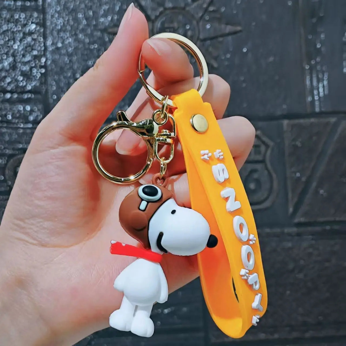 A hand holds a yellow silicone keychain featuring a white Snoopy dog with a brown floppy ear and a red collar.