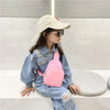 A young girl wearing a light-colored hat and sunglasses sits on a white surface with a pink nylon crossbody bag.