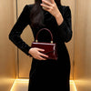 A woman holds a deep red glossy texture evening handbag against a light wood background.