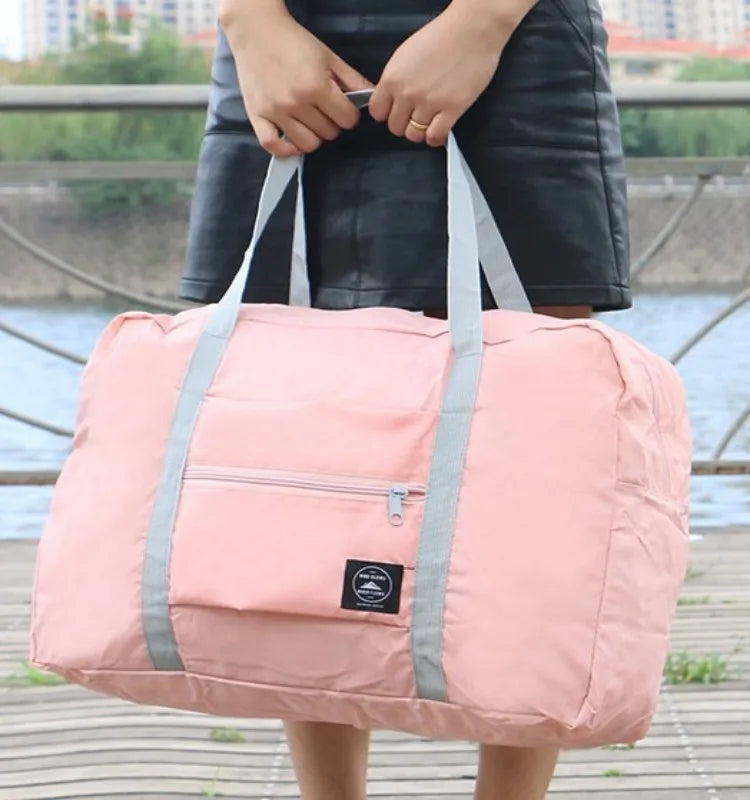 Un sac de voyage pliable rose avec des sangles grises est tenu par une personne à l'extérieur.