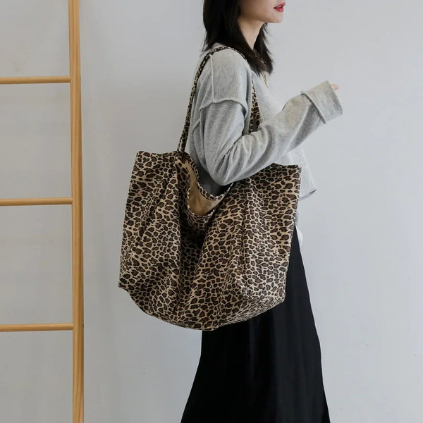 A woman holds a leopard print canvas crossbody tote bag in a light gray sweater against a white background.