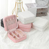 A pink compact leather jewelry travel box open to show various rings and bracelets, placed next to white storage boxes.