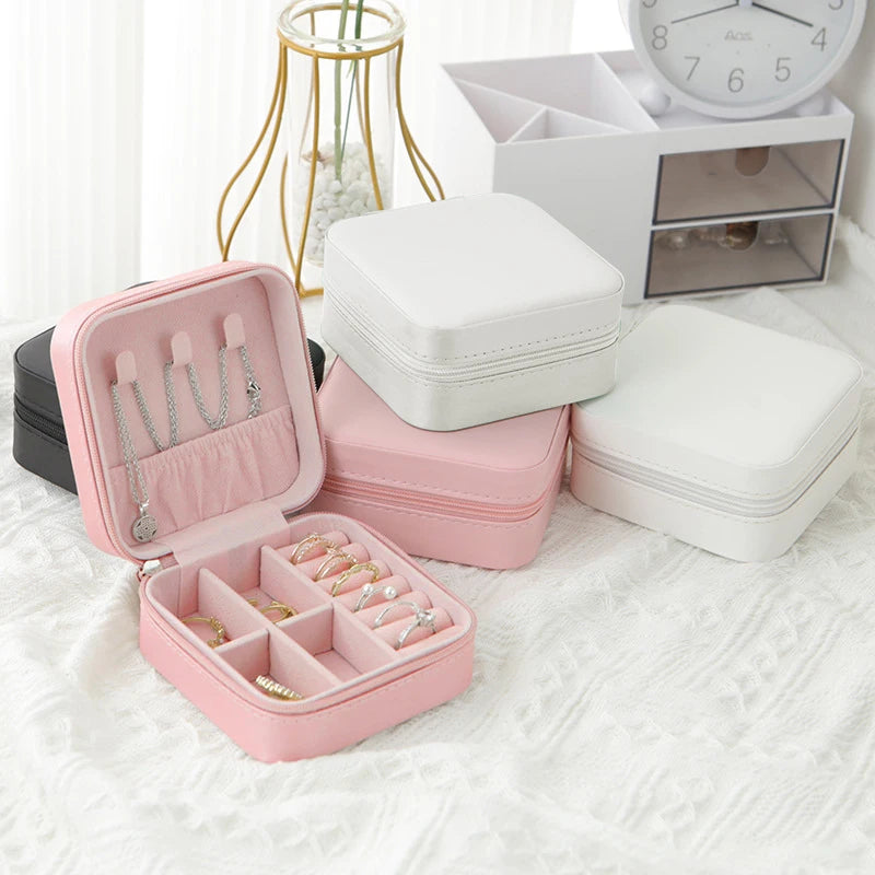 A pink compact leather jewelry travel box open to show various rings and bracelets, placed next to white storage boxes.
