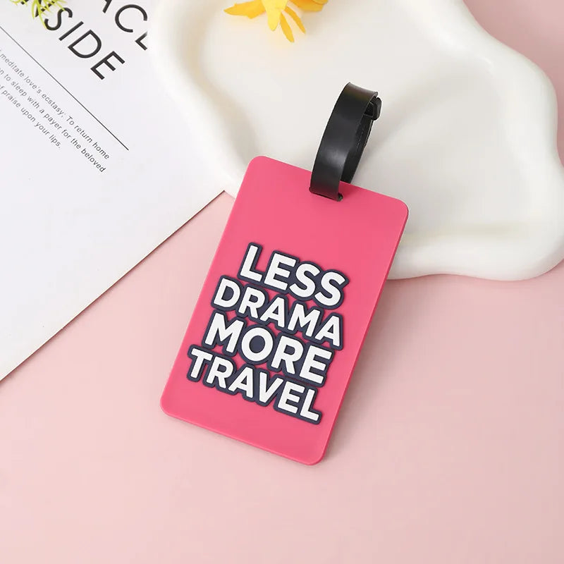 A pink silicone luggage tag with black strap and text reading Less Drama More Travel resting on a white surface.