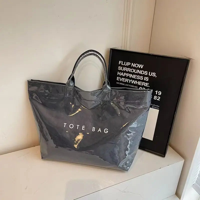 A dark gray transparent waterproof PVC tote bag is displayed on a white surface next to a framed sign.