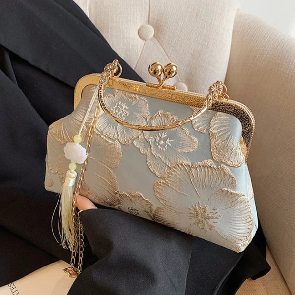 A light blue embroidered silk evening clutch with gold hardware and a chain strap is held in a black blazer.