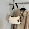 A cream colored shoulder bag with a gold chain and cherry pendant hanging on a hanger.