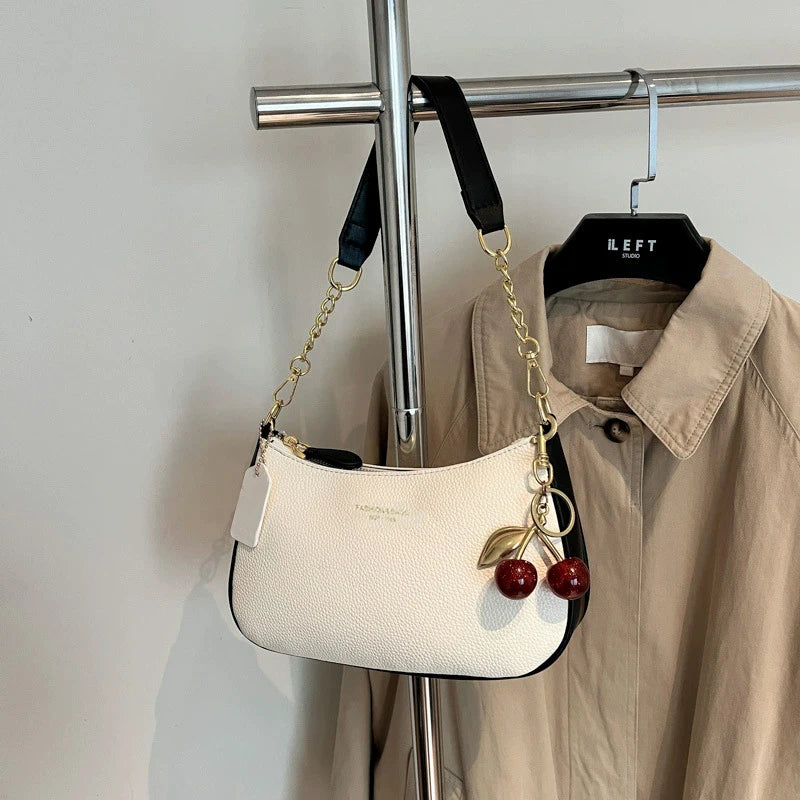 A cream colored shoulder bag with a gold chain and cherry pendant hanging on a hanger.