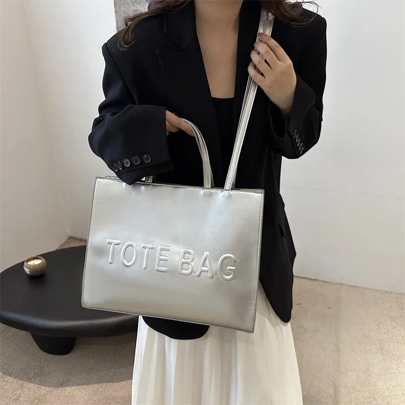 A person holds a silver tote bag with a leather strap against a white background.