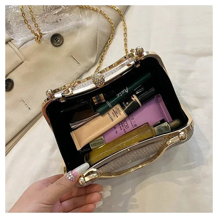 A hand holds a gold glitter crossbody evening clutch bag with a dark interior and a light beige envelope in the background.