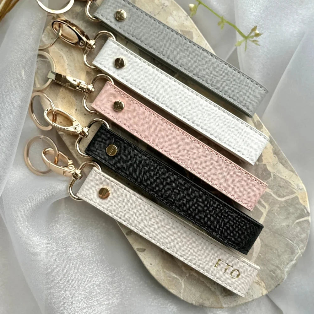 A collection of personalized leather wristlet keychains in various colors and finishes laid on a light fabric background.