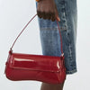 A close-up of a red PU leather shoulder bag with an adjustable strap being held by a person wearing blue jeans.
