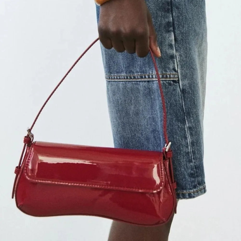 A close-up of a red PU leather shoulder bag with an adjustable strap being held by a person wearing blue jeans.