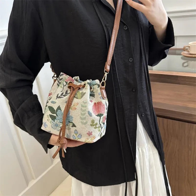 A woman holds a floral drawstring bucket bag with brown leather strap against a black shirt and white skirt.