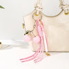 A light beige handbag with a gold chain and pink cherry charm keychain featuring braided white and pink rope.