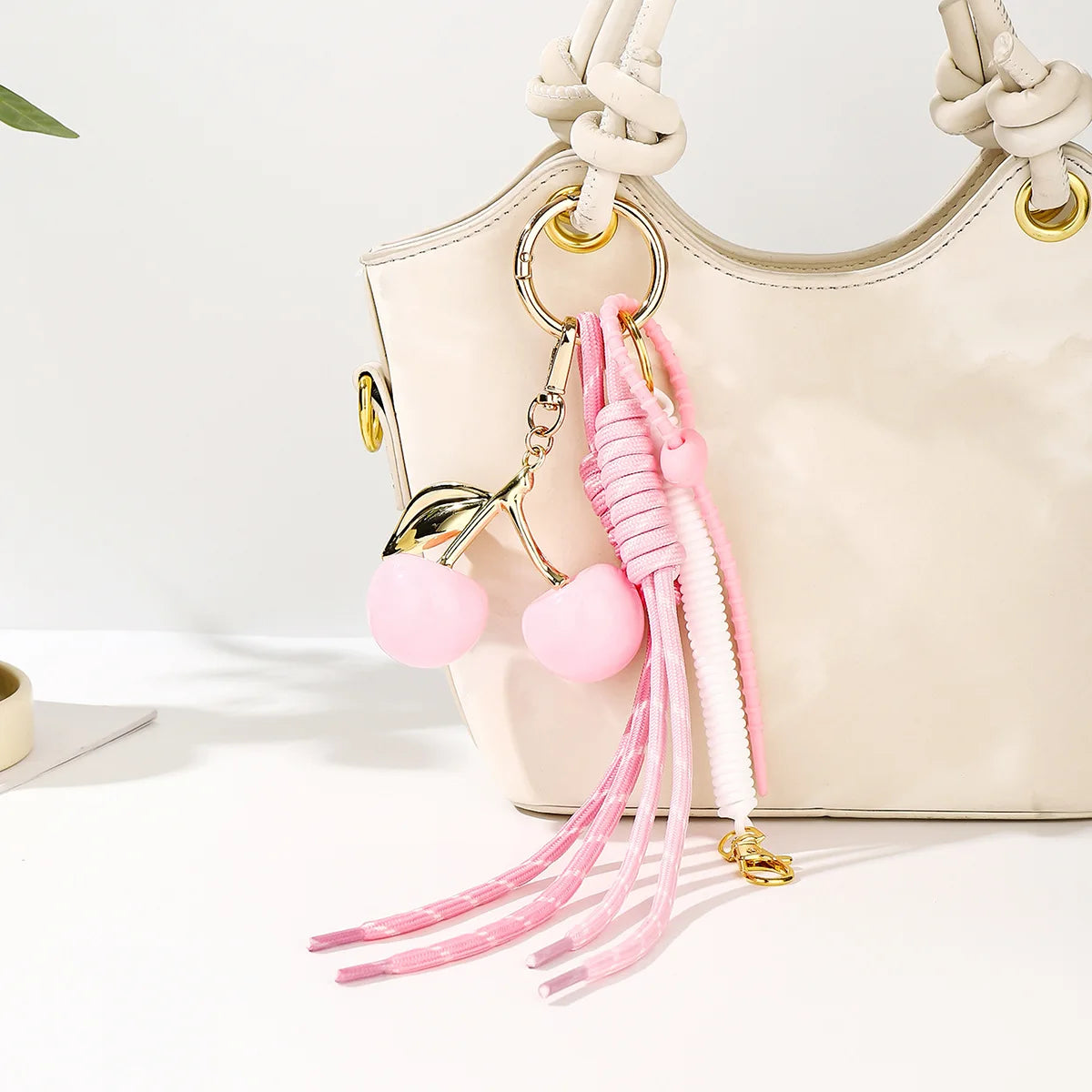 A light beige handbag with a gold chain and pink cherry charm keychain featuring braided white and pink rope.