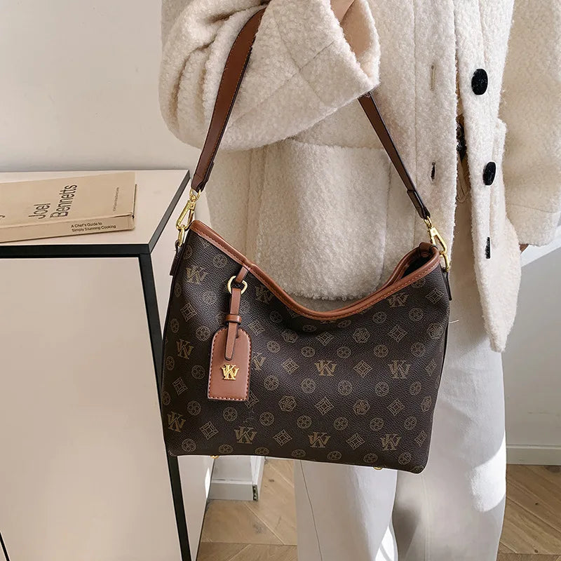 A brown leather crossbody tote bag with a gold hardware detail is shown against a white coat and a light-colored table.