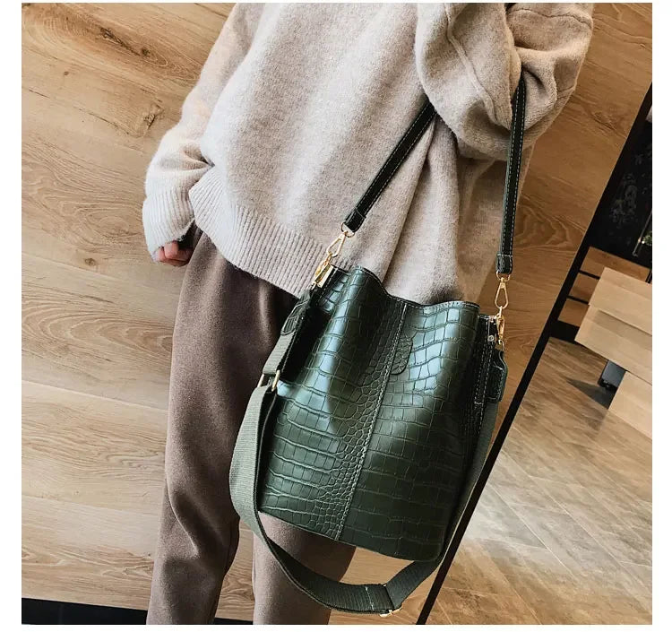 A close-up of a dark green alligator pattern leather crossbody bag carried by a person wearing a beige sweater and brown pant