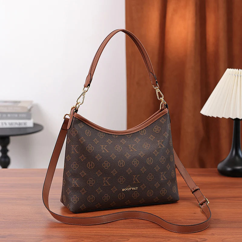 A brown leather crescent tote bag with a gold accent and a repeating letter print resting on a wooden surface.