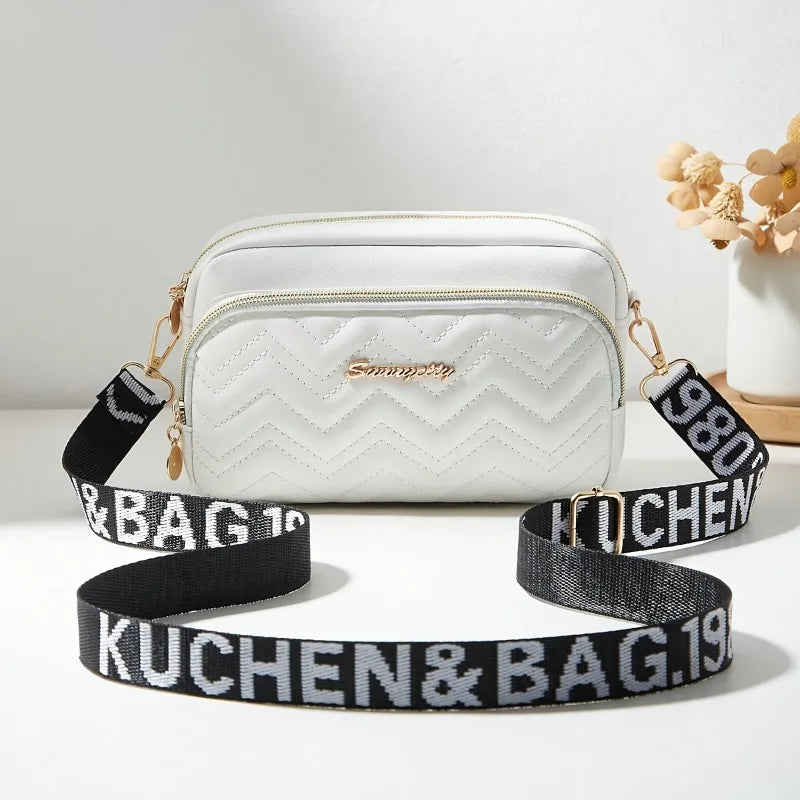 White quilted crossbody camera bag with a black and gold letter strap resting on a white surface.