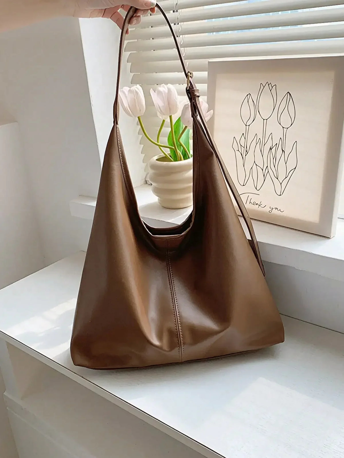 A brown PU leather slouchy hobo shoulder bag hanging on a hanger in front of a window with tulips.