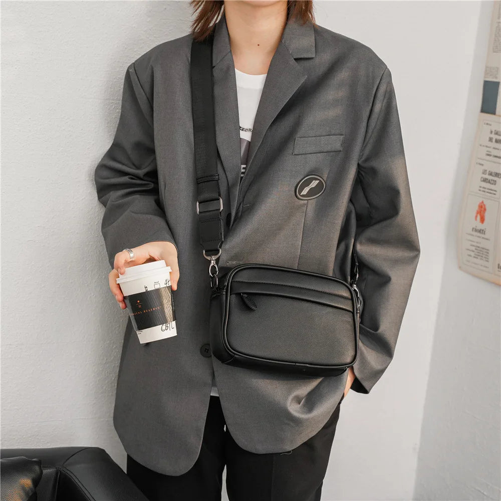 A man wearing a gray blazer and black pants carrying a black PU leather crossbody messenger bag.