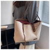 A beige canvas shoulder tote bag with brown leather accents is displayed against a light wall next to a window.