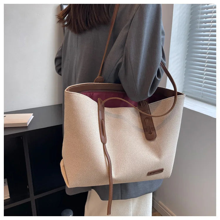 A beige canvas shoulder tote bag with brown leather accents is displayed against a light wall next to a window.